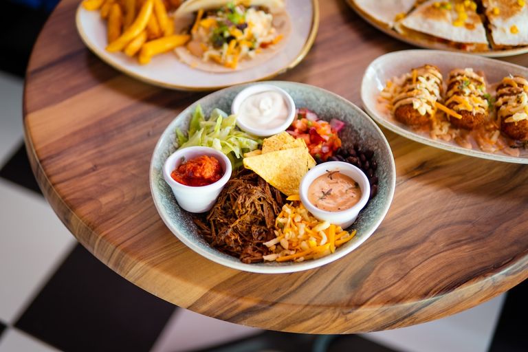 A colorful Mexican bowl with shredded beef, cheese, chips, and small dip cups on a wooden table alongside other dishes at Rosa Mexicano.